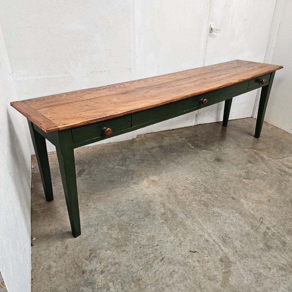 A long French painted pine console table featuring a natural wood plank top, a dark green painted base with tapered square legs, and three inline drawers with wooden knobs.