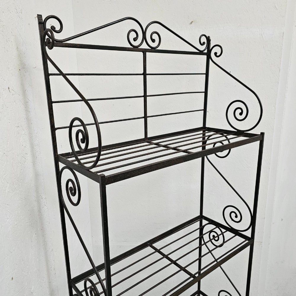 A tall dark wrought iron bakers rack featuring four slatted open shelves, side lattice supports, and decorative scrollwork along the upper pediment. View II