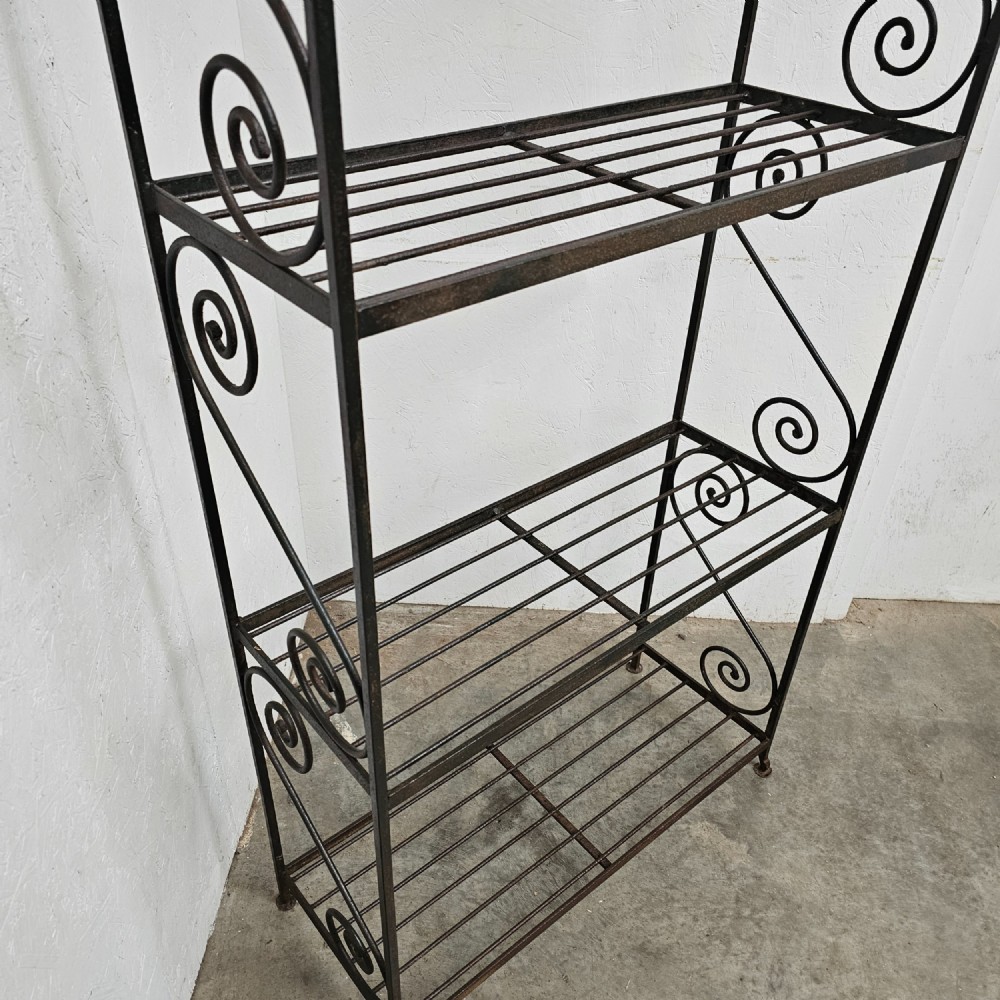 A tall dark wrought iron bakers rack featuring four slatted open shelves, side lattice supports, and decorative scrollwork along the upper pediment. View III