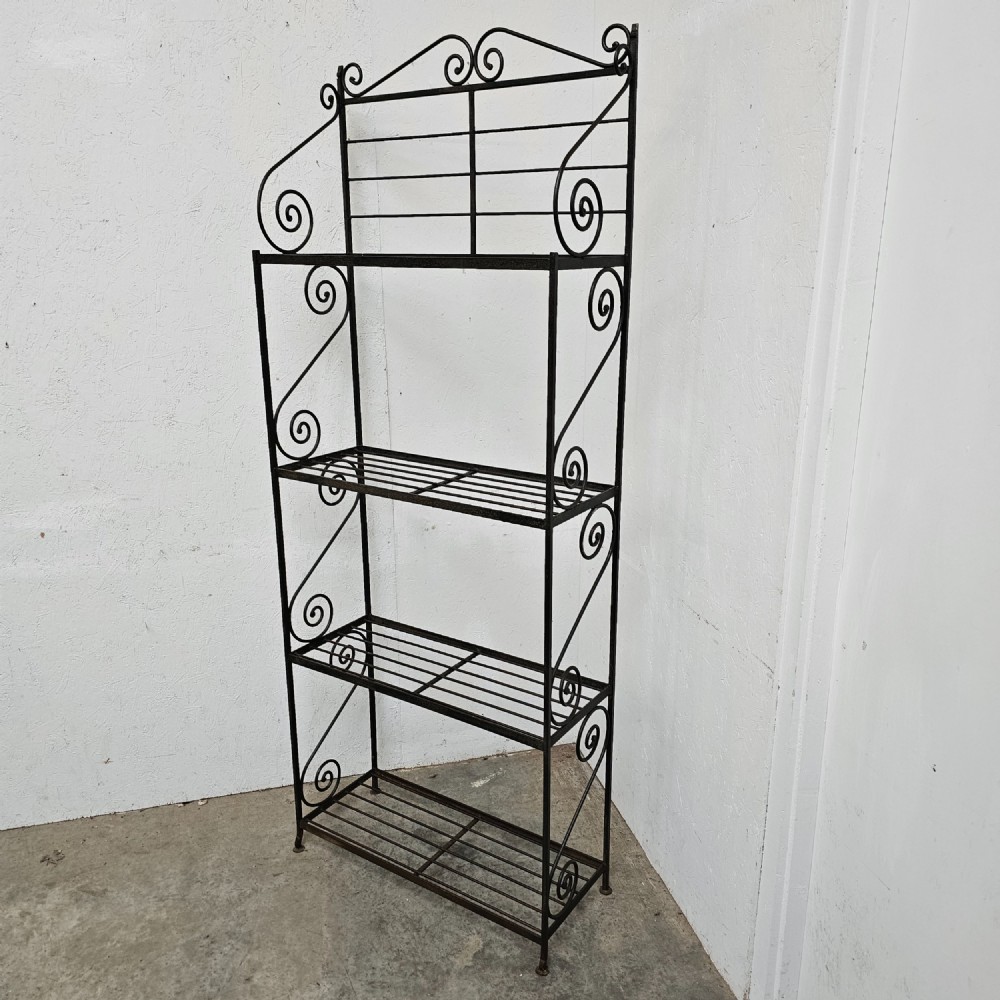 A tall dark wrought iron bakers rack featuring four slatted open shelves, side lattice supports, and decorative scrollwork along the upper pediment. View V