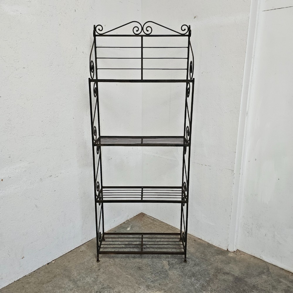 A tall dark wrought iron bakers rack featuring four slatted open shelves, side lattice supports, and decorative scrollwork along the upper pediment.