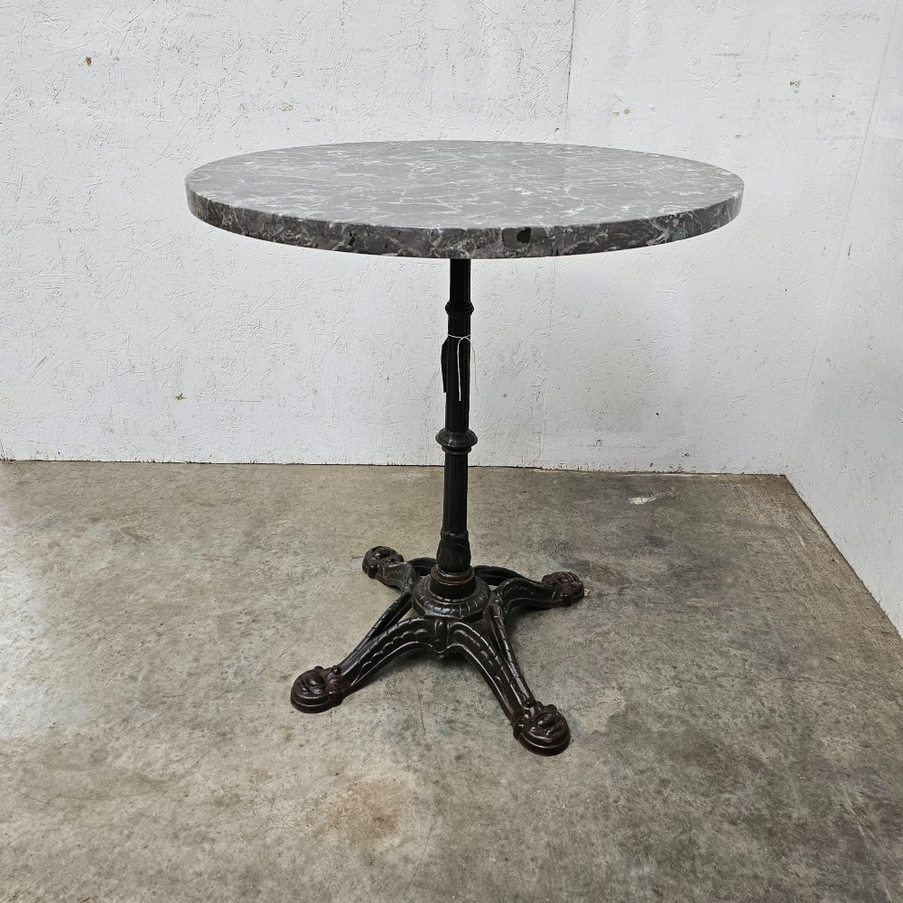 A French bistro table featuring a circular grey veined marble top resting on a dark cast iron pedestal base with decorative feet.