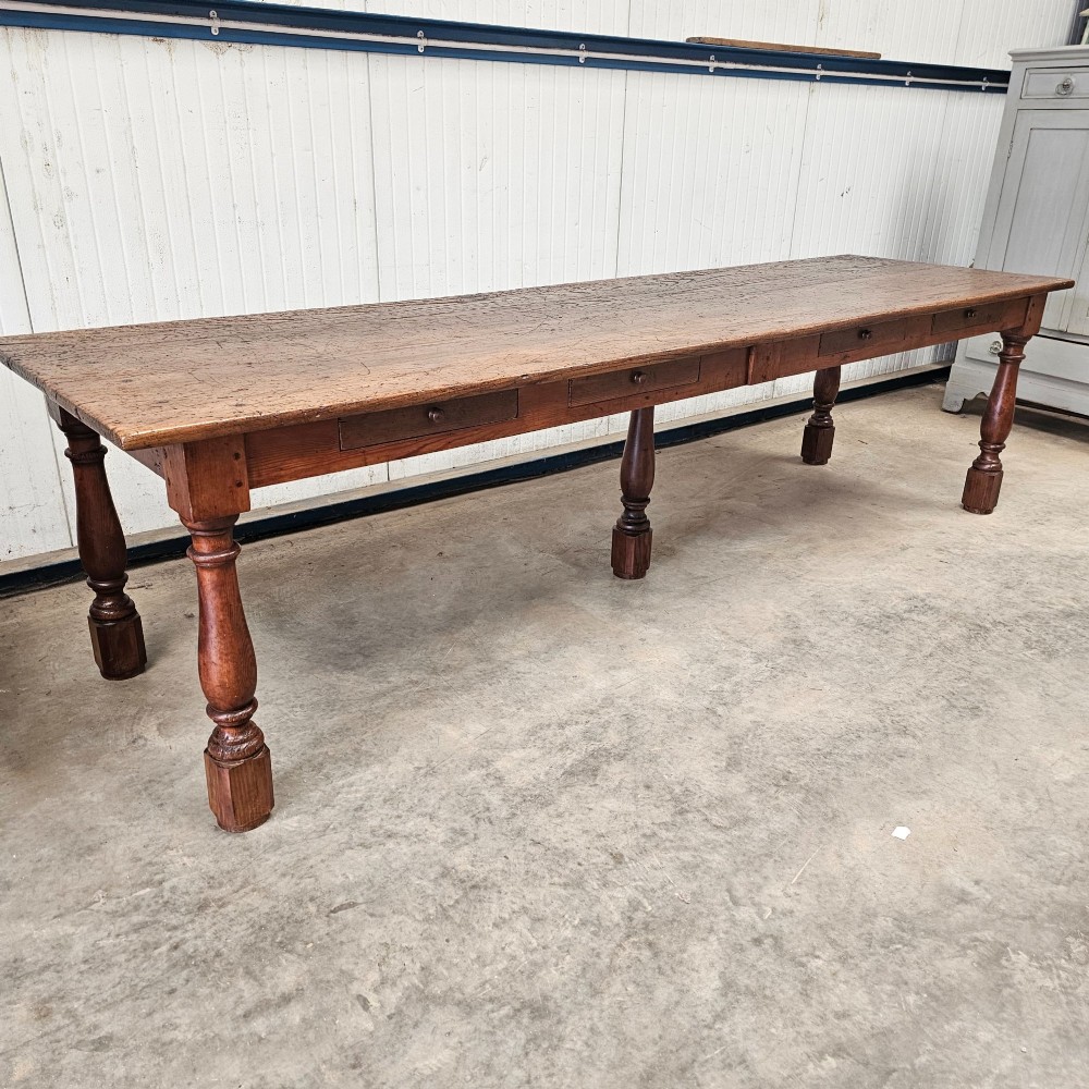 A long rustic chestnut and pine farmhouse dining table featuring six turned legs and multiple side drawers along the wooden frame.