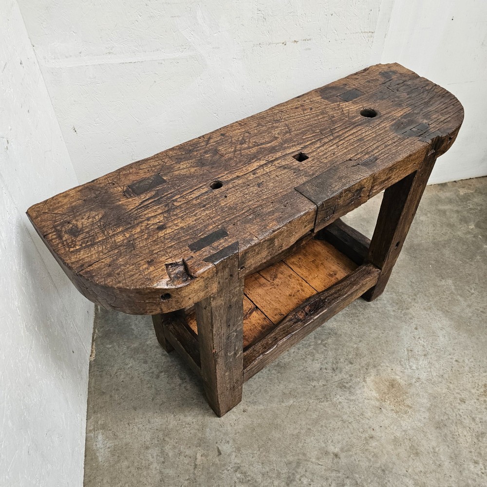 A small primitive French elm workbench featuring a thick distressed wood top, heavy trestle legs, and a lower storage shelf. View V