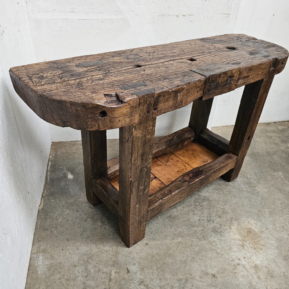 A small primitive French elm workbench featuring a thick distressed wood top, heavy trestle legs, and a lower storage shelf. View VI