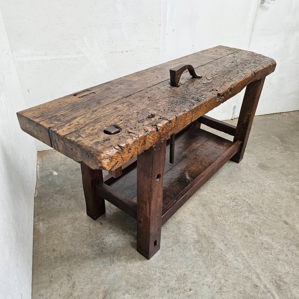 A rustic antique French dark oak workbench featuring a thick timber top slab, lower shelf, sturdy block legs, and an integrated forged iron clamping bar. View II