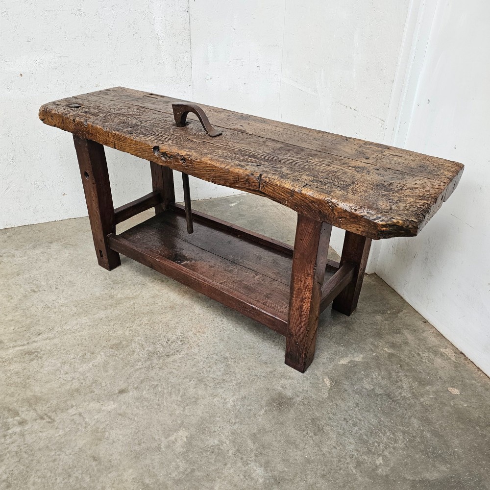 A rustic antique French dark oak workbench featuring a thick timber top slab, lower shelf, sturdy block legs, and an integrated forged iron clamping bar. View III