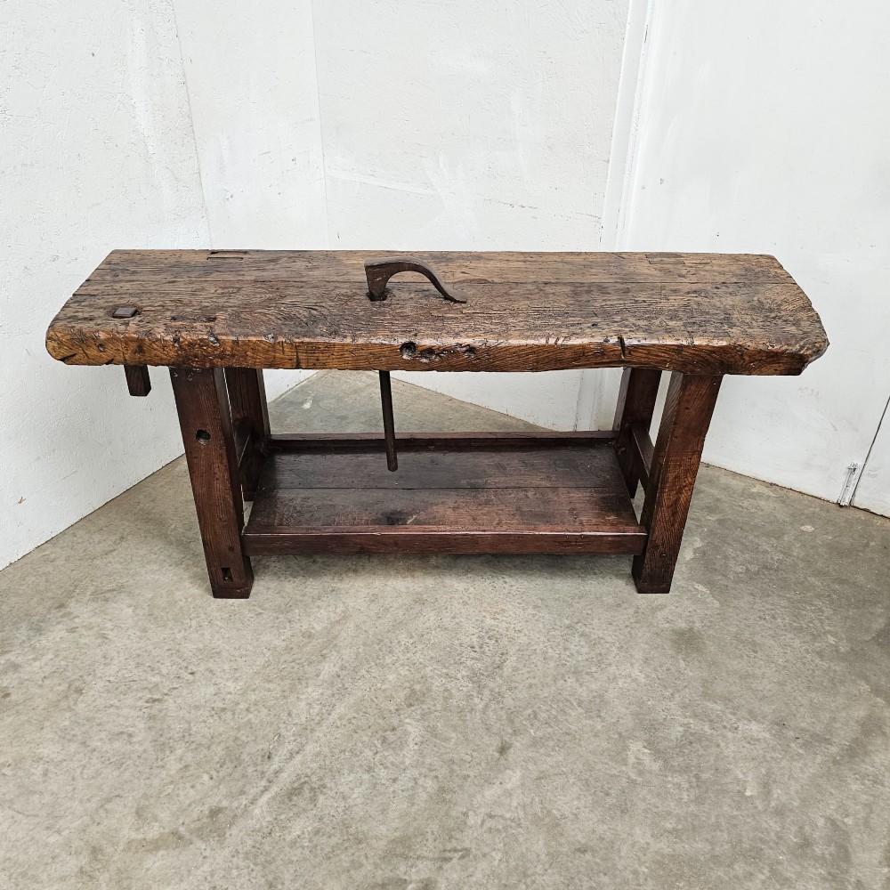A rustic antique French dark oak workbench featuring a thick timber top slab, lower shelf, sturdy block legs, and an integrated forged iron clamping bar. View IV