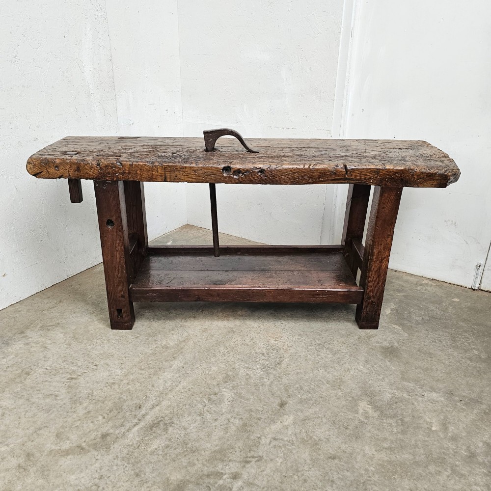 A rustic antique French dark oak workbench featuring a thick timber top slab, lower shelf, sturdy block legs, and an integrated forged iron clamping bar.