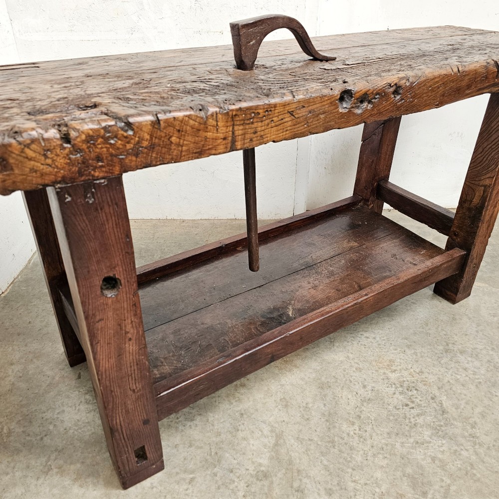 A rustic antique French dark oak workbench featuring a thick timber top slab, lower shelf, sturdy block legs, and an integrated forged iron clamping bar. View V