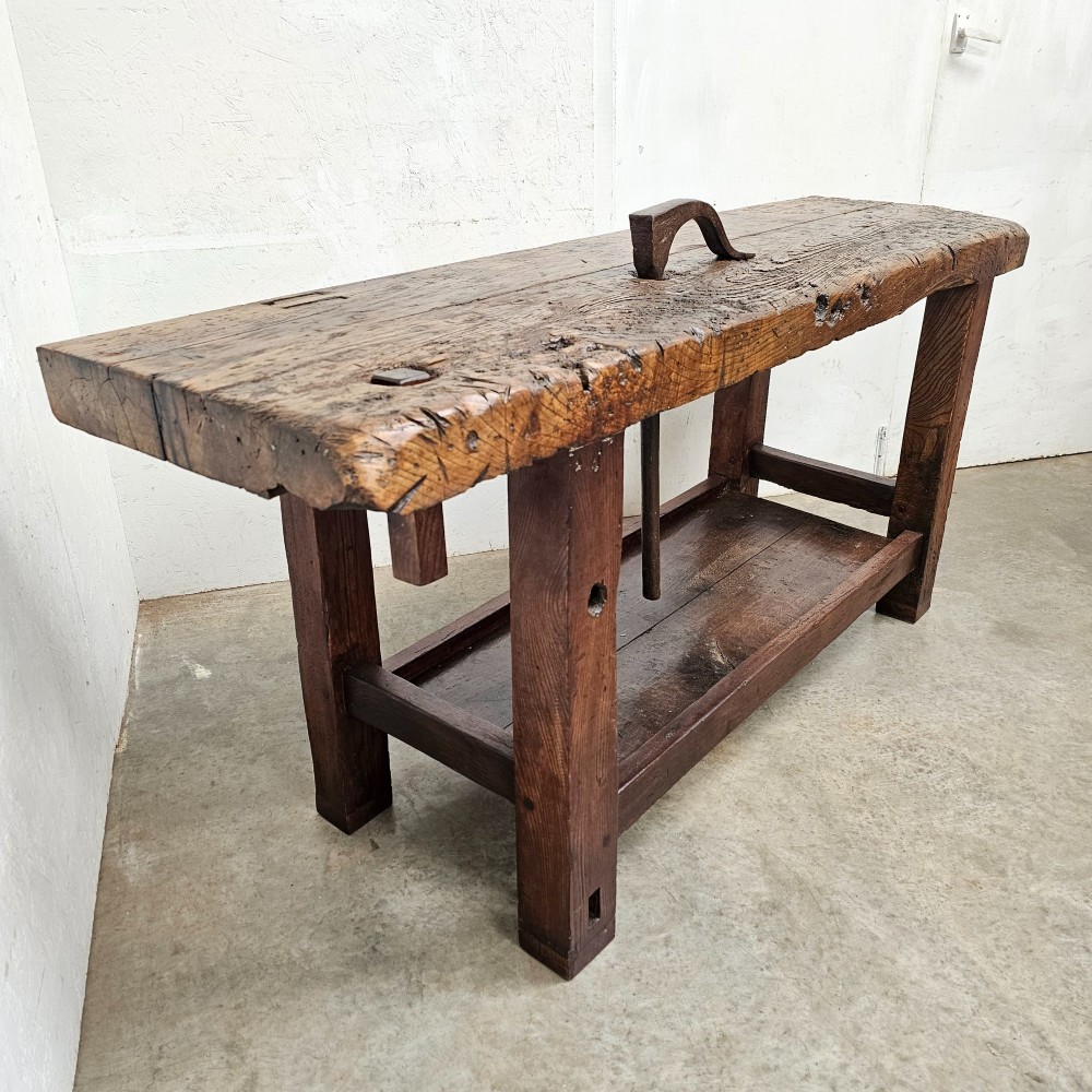 A rustic antique French dark oak workbench featuring a thick timber top slab, lower shelf, sturdy block legs, and an integrated forged iron clamping bar. View VI