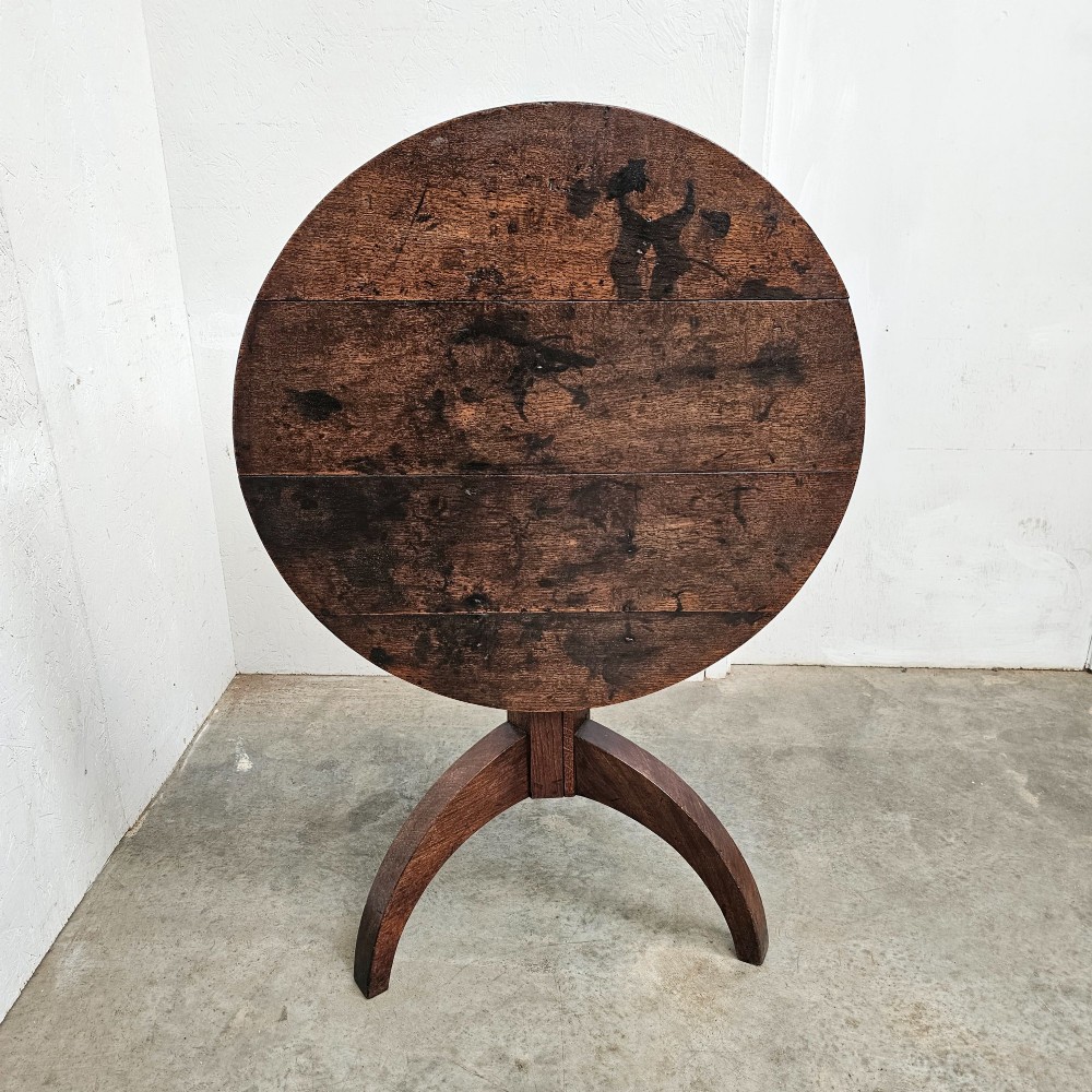 A dark oak round tilt-top table shown tilted vertically, supported by a central column and three curved tripod legs.