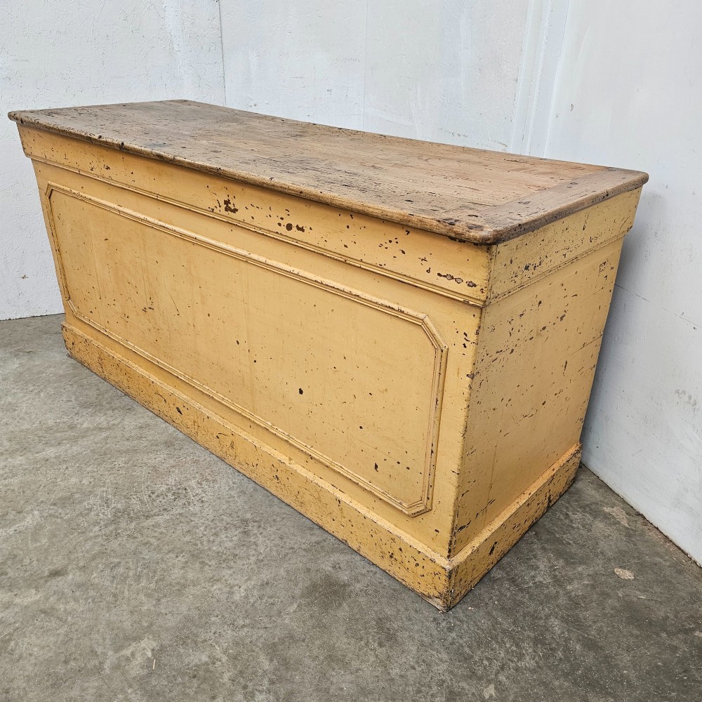 A large rectangular timber shop counter featuring a distressed yellow painted frame with a moulded front panel and a natural wood top. View II