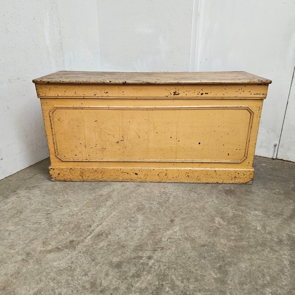 A large rectangular timber shop counter featuring a distressed yellow painted frame with a moulded front panel and a natural wood top.