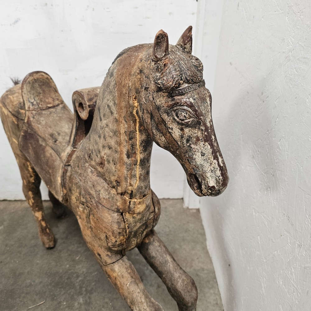 A 19th-century French hand-carved wooden carousel horse featuring a prancing pose, carved saddle, black painted mane, and weathered grey-brown timber finish. View V