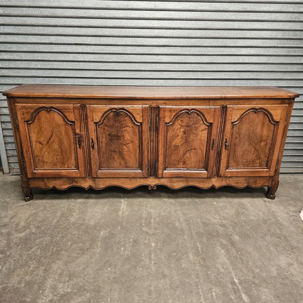 An 18th-century French provincial walnut four-door sideboard featuring hand-carved arched door panels, dark metal escutcheons, and a scalloped lower apron.