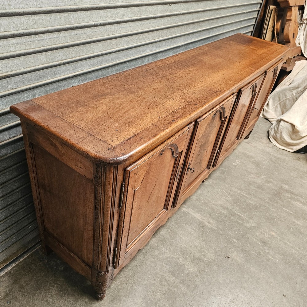 An 18th-century French provincial walnut four-door sideboard featuring hand-carved arched door panels, dark metal escutcheons, and a scalloped lower apron. View V