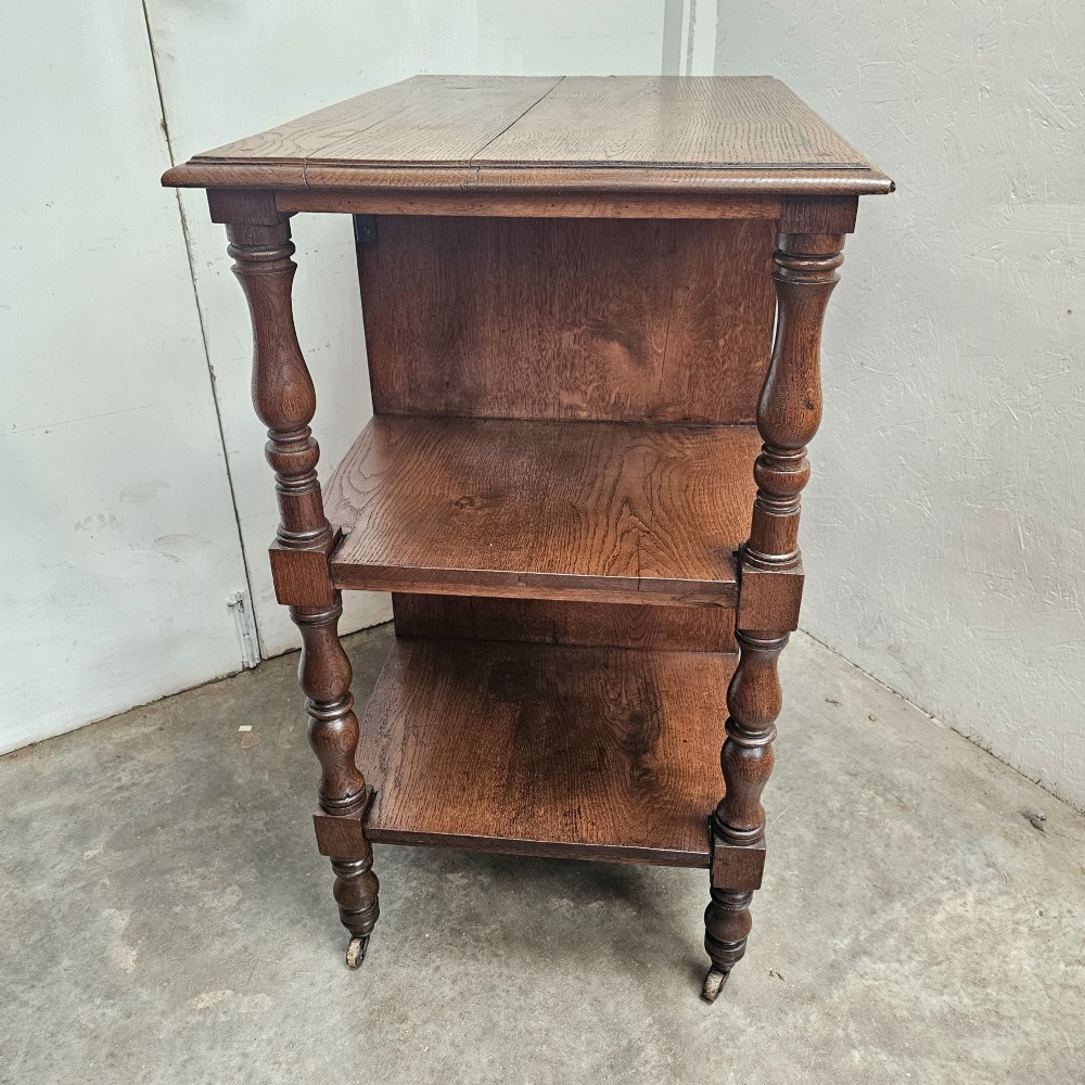 A dark oak library cart featuring three tiered shelves, central partitions, turned legs, and small metal castors. View II