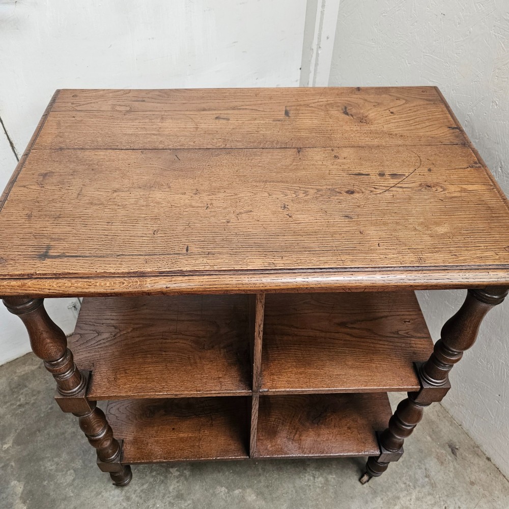A dark oak library cart featuring three tiered shelves, central partitions, turned legs, and small metal castors. View III