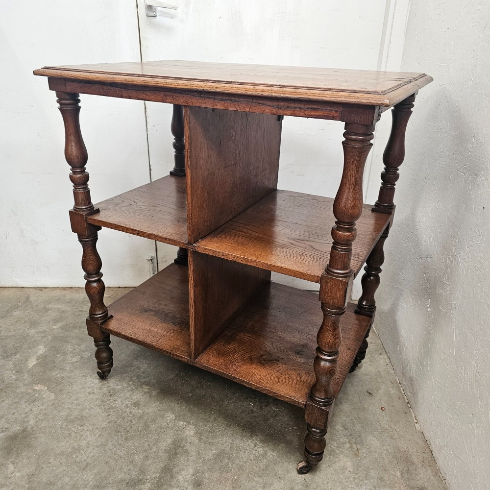 A dark oak library cart featuring three tiered shelves, central partitions, turned legs, and small metal castors.