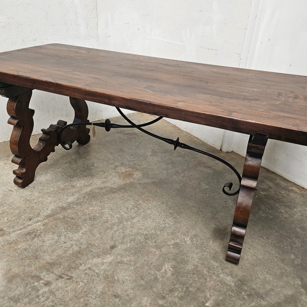 A dark brown rectangular wooden dining table with carved trestle legs connected by curved wrought iron stretchers. View III