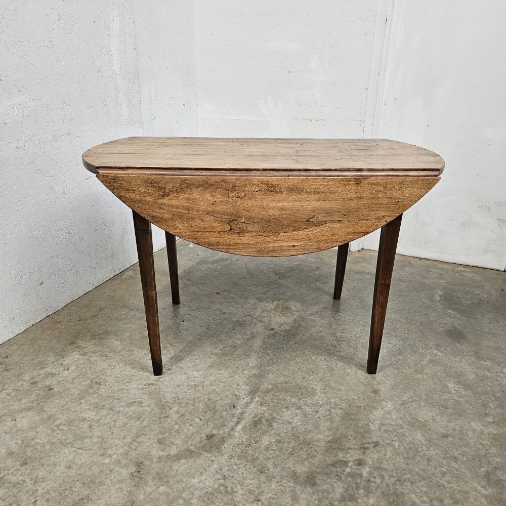 An antique wooden French drop-leaf table with semi-circular side leaves folded down and four tapered wooden legs.