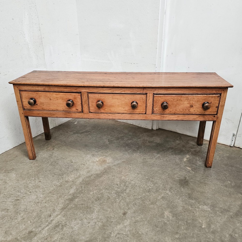 An English country pine and elm dresser base featuring a planked top, three frieze drawers with turned knobs, and four square tapered legs.