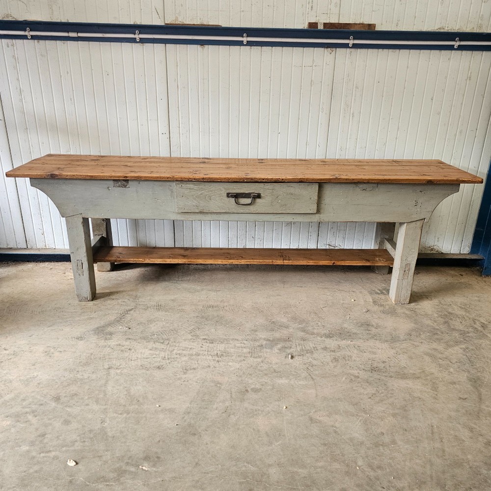 A long rustic pine worktable featuring a natural wood top, a light grey painted frame with a single drawer, and a lower shelf.