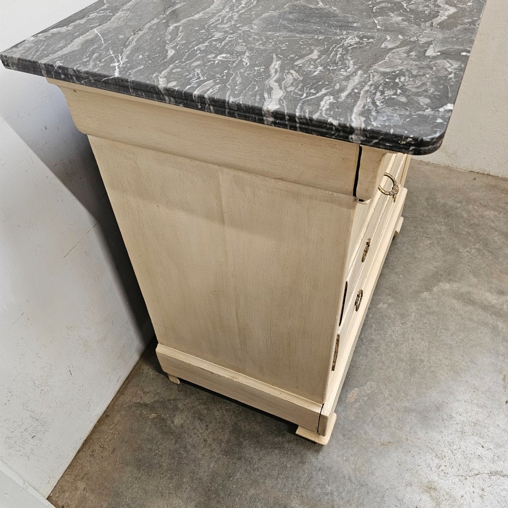 An off-white painted Louis Philippe chest of drawers with a dark marble top and brass ring handles. View VI