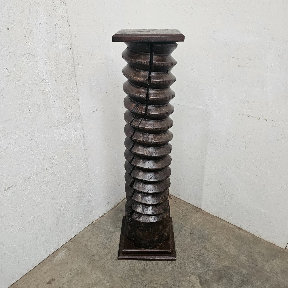 A tall hand-carved dark timber wine press screw thread with a square top and base, repurposed as a sculptural display pedestal.