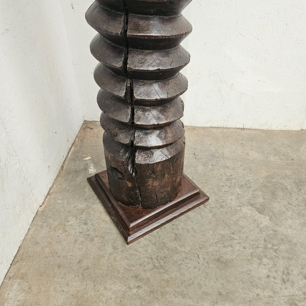 A tall hand-carved dark timber wine press screw thread with a square top and base, repurposed as a sculptural display pedestal. View III