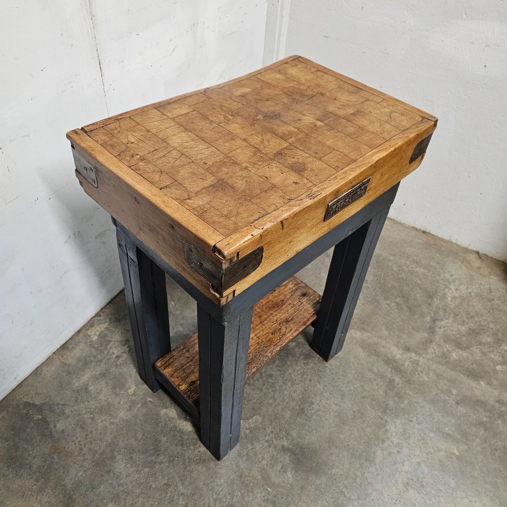 A small antique French butchers block featuring an end-grain beech top with a brass makers plate set on a dark painted wooden stand with a lower shelf. View II