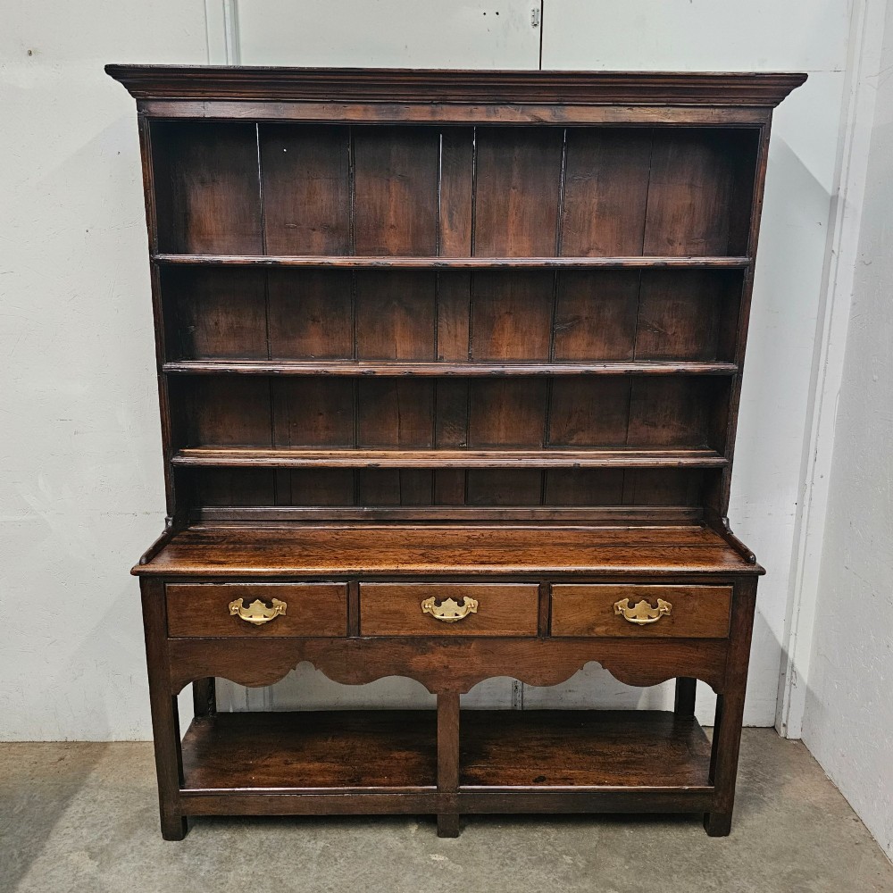 An antique dark oak potboard dresser featuring a tall open tiered plate rack over three drawer units with brass handles and a low open display shelf.