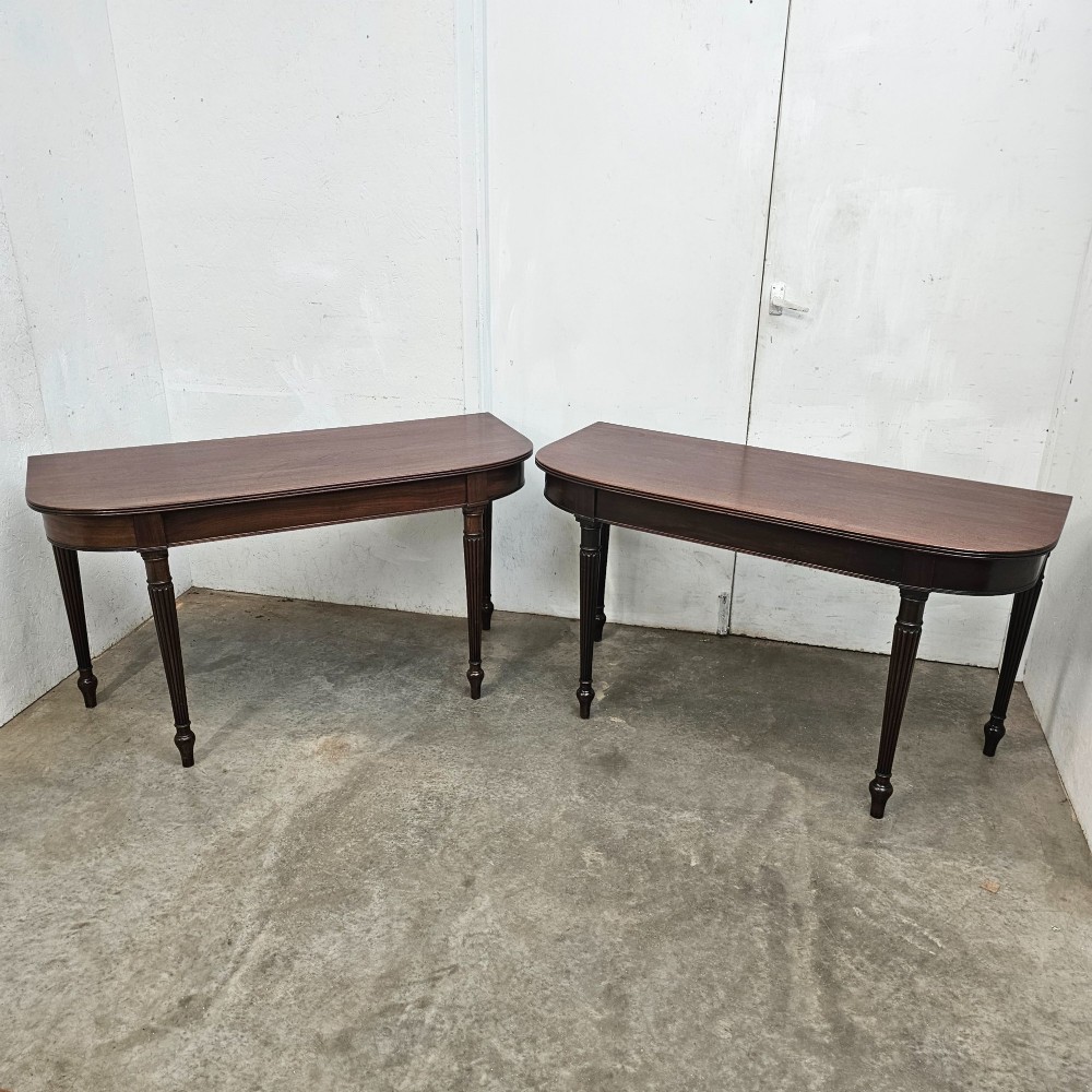 A pair of English mahogany console tables featuring D-shaped tops, subtle reeded aprons, and slender fluted tapered legs.