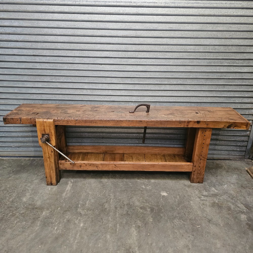 A long, heavy French oak workbench with an integrated wooden vice, lower storage shelf, and aged iron hardware.