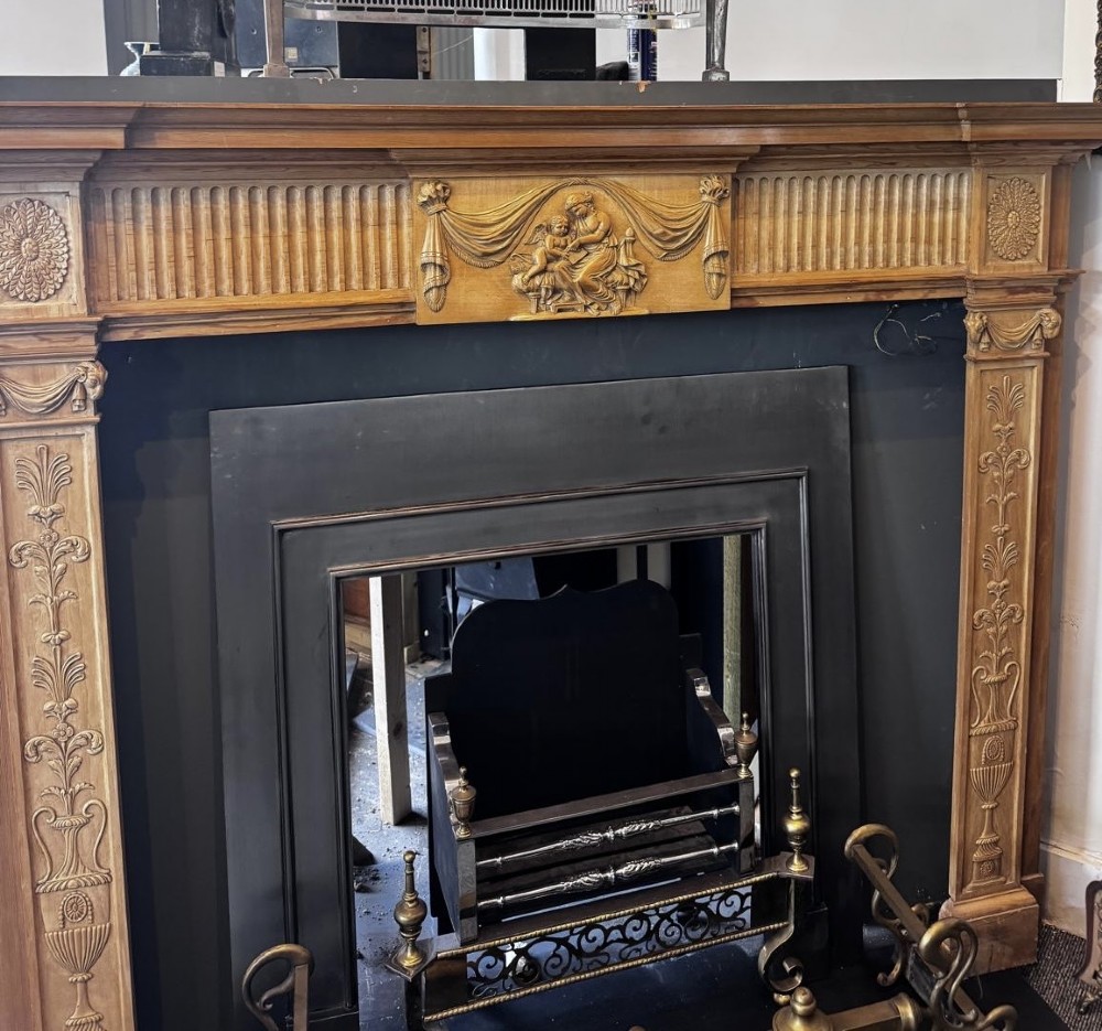 A Georgian carved wooden fireplace surround featuring a central figural relief tablet, fluted frieze, drapery swags, and classical pilasters.