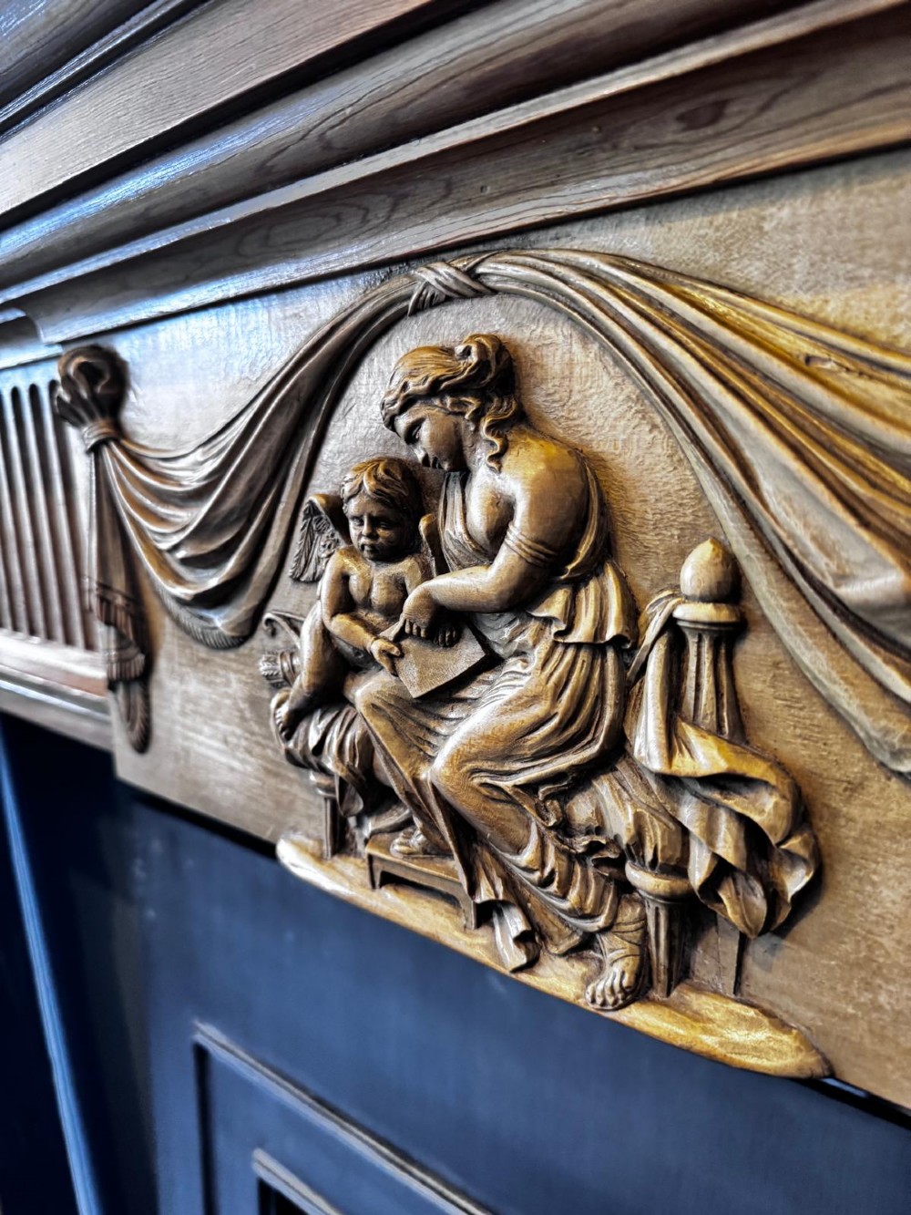 A Georgian carved wooden fireplace surround featuring a central figural relief tablet, fluted frieze, drapery swags, and classical pilasters. View V