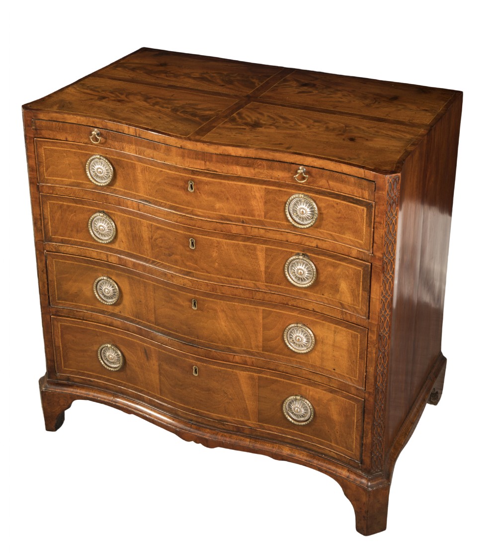 A George III serpentine-front chest of drawers featuring four graduated drawers with brass ring handles and carved blind fretwork on chamfered corners.