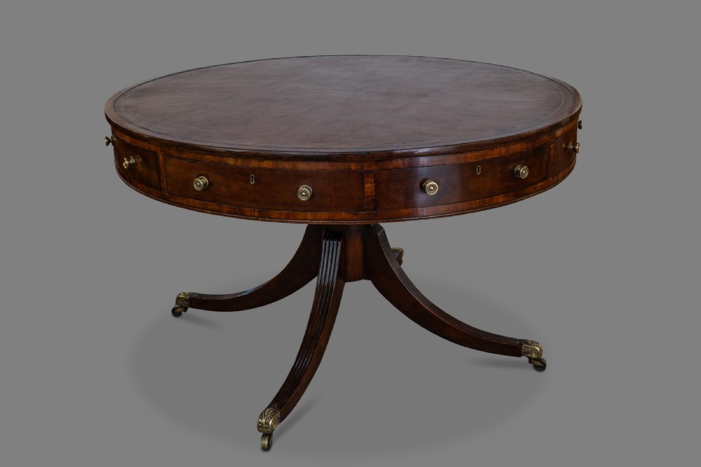 A circular George III mahogany drum table featuring a dark leather top, perimeter drawers with brass knobs, and a central pedestal with four reeded legs on brass castors.