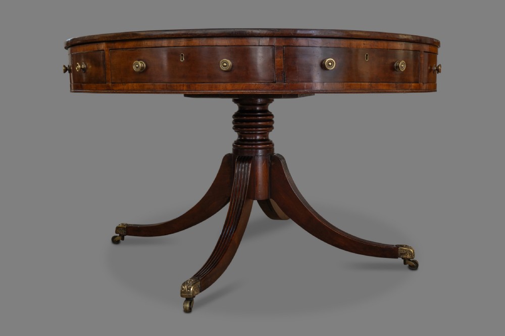 A circular George III mahogany drum table featuring a dark leather top, perimeter drawers with brass knobs, and a central pedestal with four reeded legs on brass castors. View II