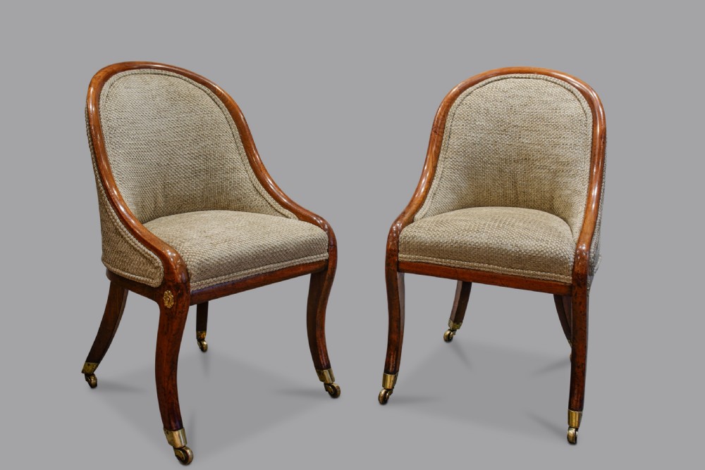 A pair of low-backed Regency mahogany slipper chairs featuring curved wooden frames, upholstered seats and backs, and sabre legs with brass castors.