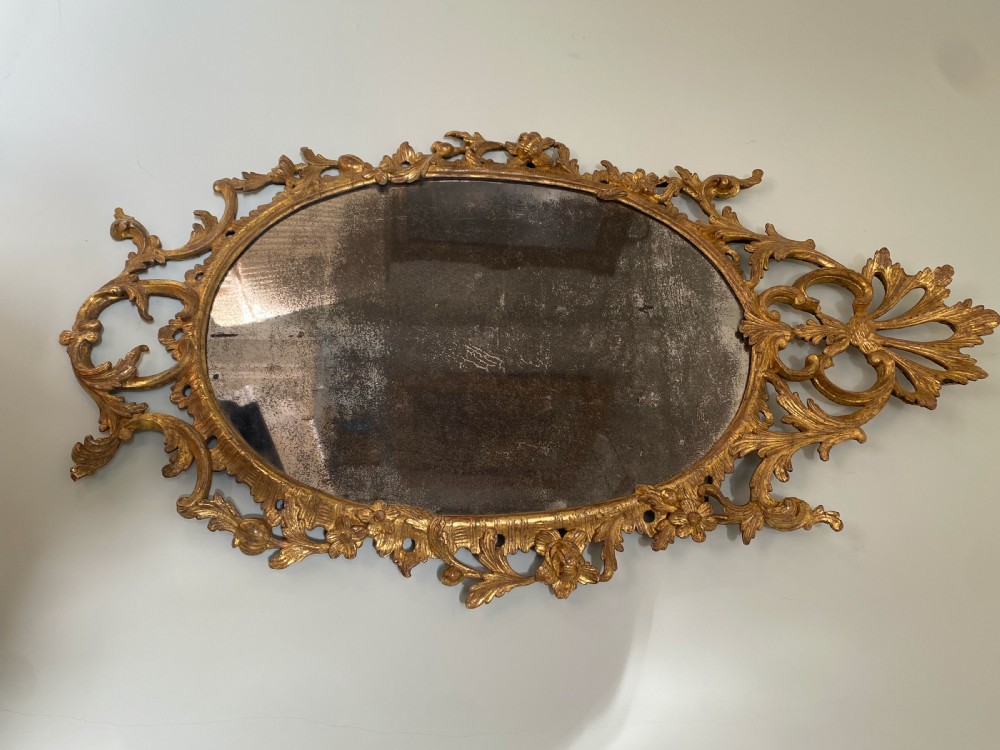 An oval George III carved giltwood wall mirror featuring pierced acanthus leaf cresting and scrollwork surrounding aged mirror glass. View II
