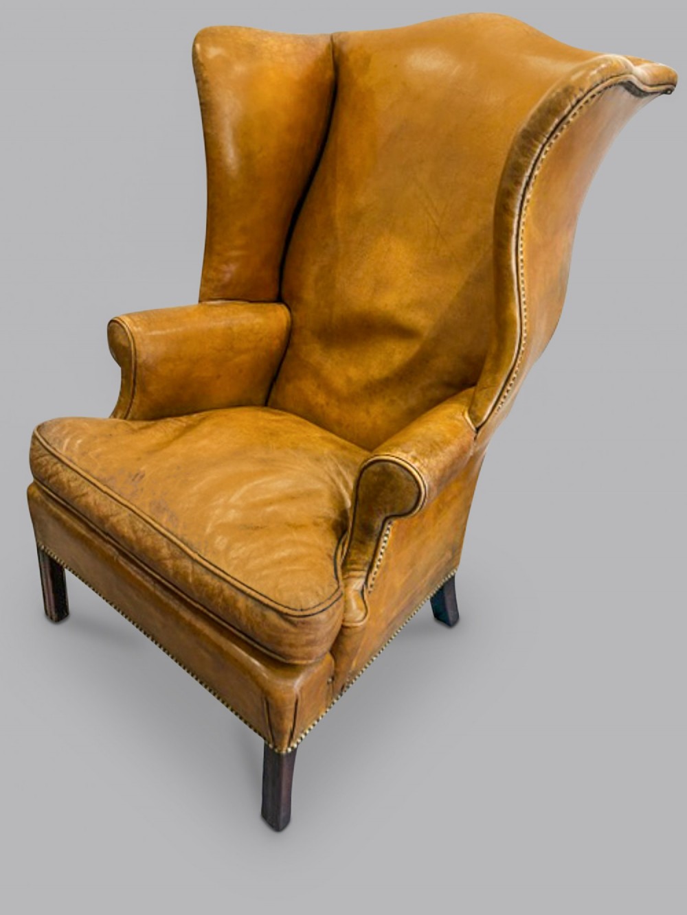 A George III wingback armchair upholstered in tan leather with brass stud detail along the seams and dark wood legs.
