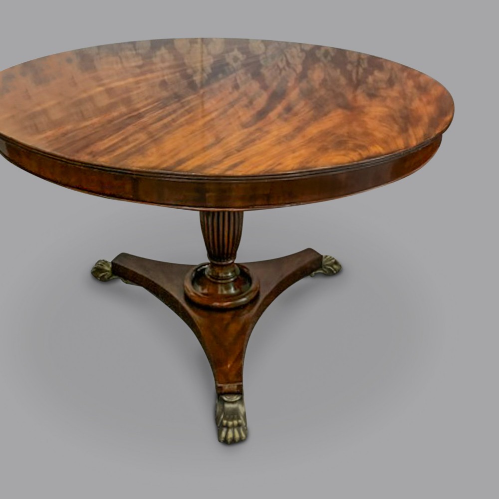A circular George III mahogany centre table featuring a polished flame mahogany top, a fluted turned pedestal, and a triform base resting on brass claw feet.