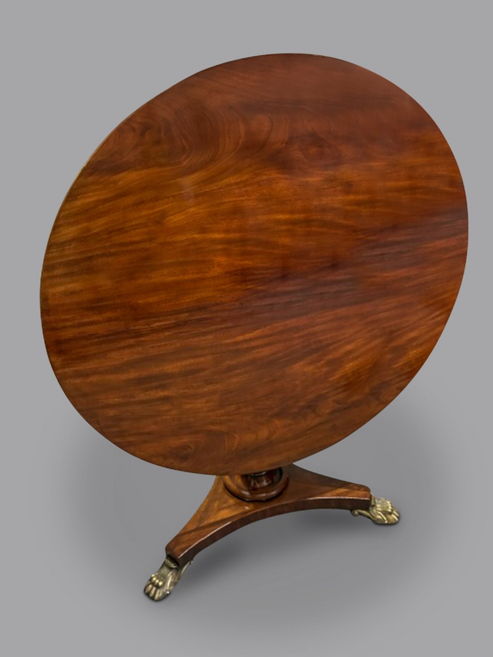 A circular George III mahogany centre table featuring a polished flame mahogany top, a fluted turned pedestal, and a triform base resting on brass claw feet. View II