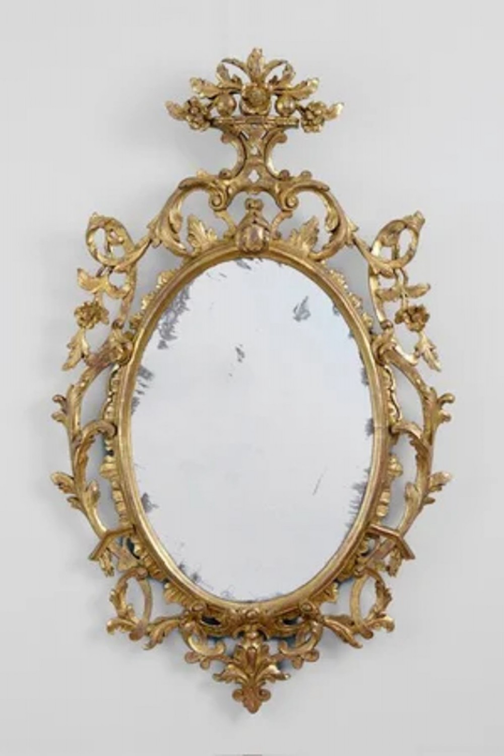 A George III oval wall mirror framed in carved giltwood with intricate openwork foliate scrolls and a floral cresting.