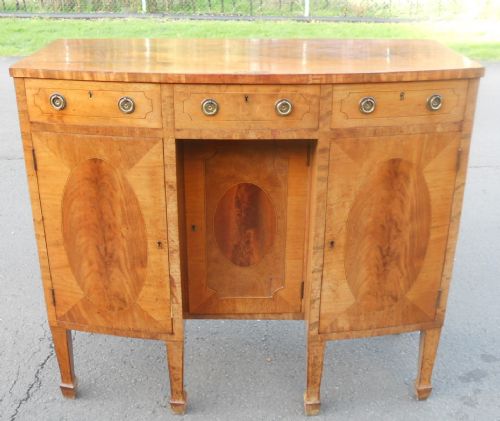 fig 1. A small bowfront mahogany sideboard featuring crossbanded drawer fronts, oval veneer cabinet door inlays, brass ring pull handles, and square tapered legs.