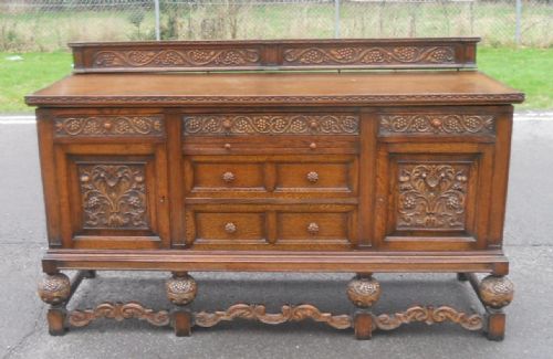 fig 1. A large carved oak sideboard featuring a low gallery back, central drawers, flanking cupboard doors with foliate carvings, and carved bulbous feet.