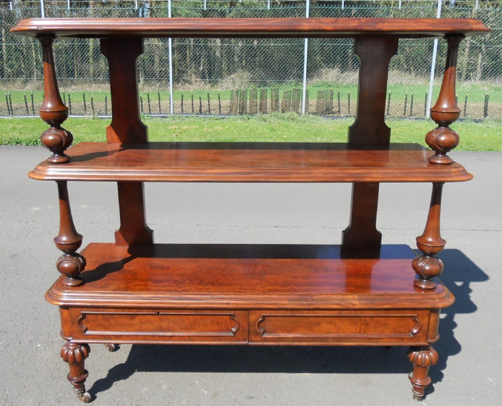 A Victorian three-tier mahogany dumb waiter featuring turned baluster supports, lower moulded drawers, and carved legs on brass castors.