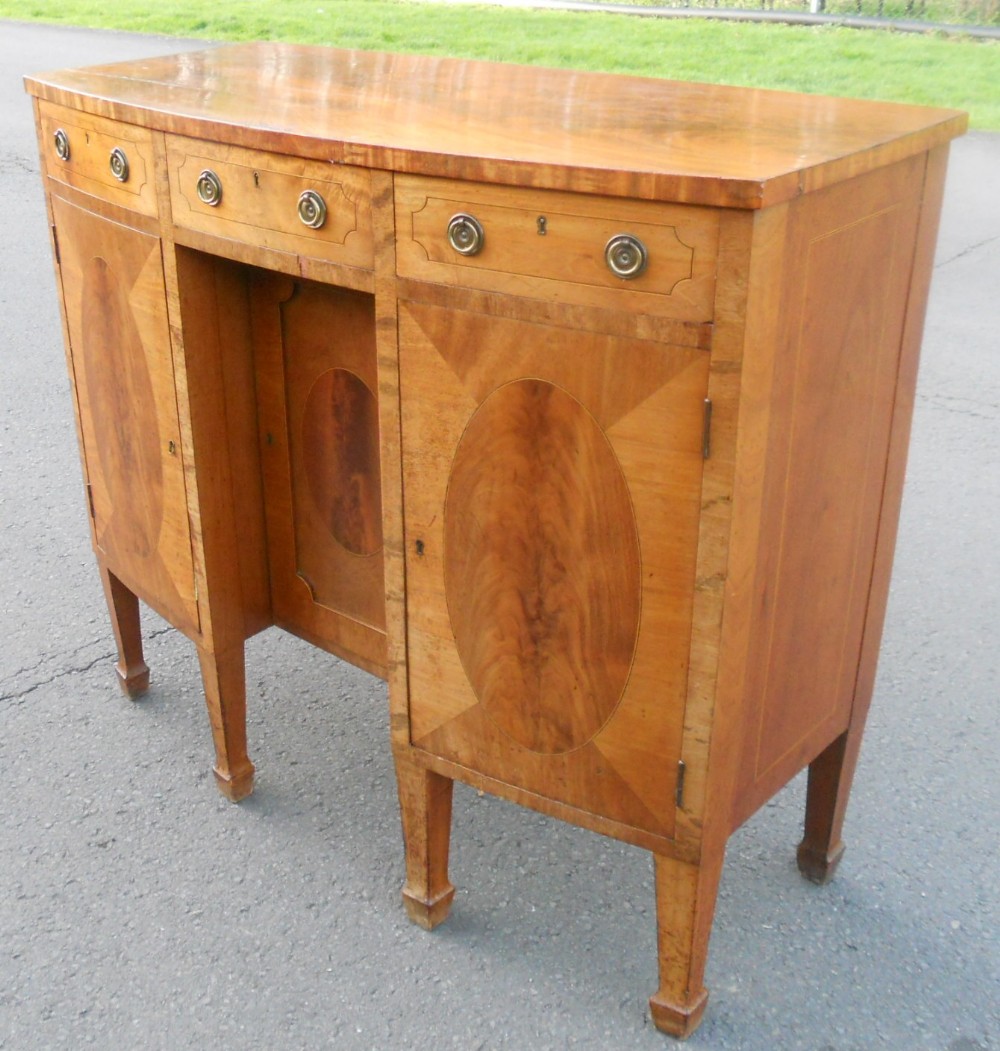 A small bowfront mahogany sideboard featuring crossbanded drawer fronts, oval veneer cabinet door inlays, brass ring pull handles, and square tapered legs.